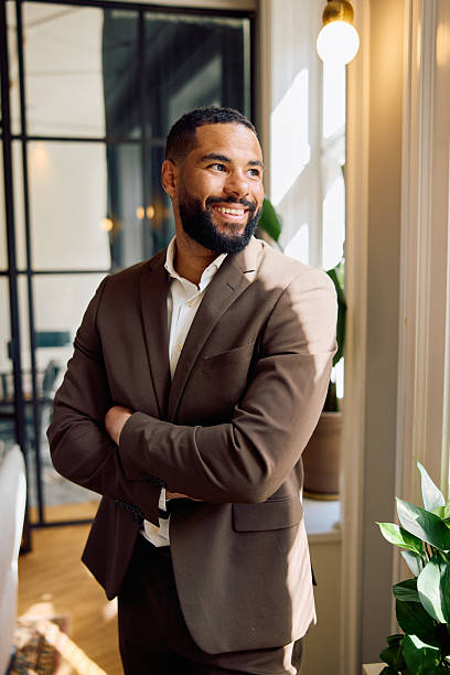 istock Pensive professional man smiling confidently while standing near brightly lit office window with plants 2234648105
