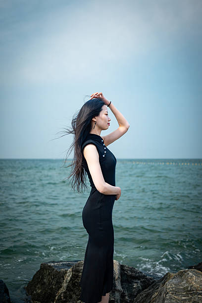 A woman standing by the water's edge, looking out at the sea. She is wearing a black dress and has her hands clasped behind her back. The calm ocean and overcast sky create a contemplative mood, highlighting a moment of reflection and tranquility