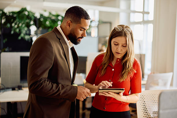 Focused man in a brown suit and woman in a red blouse standing side by side at a desk, reviewing information on a tablet in a bright modern office setting.