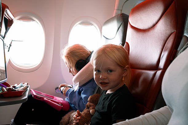 A waist-up shot of two sisters sitting next to each other on a commercial aeroplane. The girl furthest away has an iPad and a neck support. The girl nearest is cuddling a teddy bear. In flight