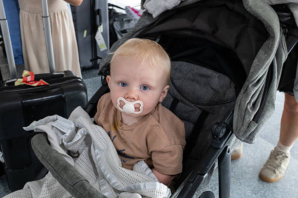 A full shot of a baby sitting in a pram with a pacifier in his mouth looking into he camera. Unrecognisable family members are standing nearby. Located in Northumberland, England.