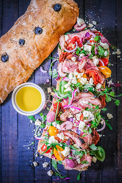 A delectable open-faced sandwich featuring colorful ingredients including ripe tomatoes, arugula, feta cheese, and olives on rustic bread. Accompanied by olive oil for dipping, the spread exudes Mediterranean flavors on a dark wood background, offering excellent character and warmth for culinary presentations.