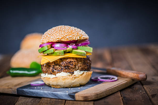 A close-up of a mouthwatering cheeseburger styled with avocado slices, red onion, and cheddar cheese, served on a sesame seed bun. Presented on a rustic wooden surface, this image captures the essence of artisanal cuisine. Vibrant colors and appetizing textures make it ideal for showcasing gourmet food and dining concepts.