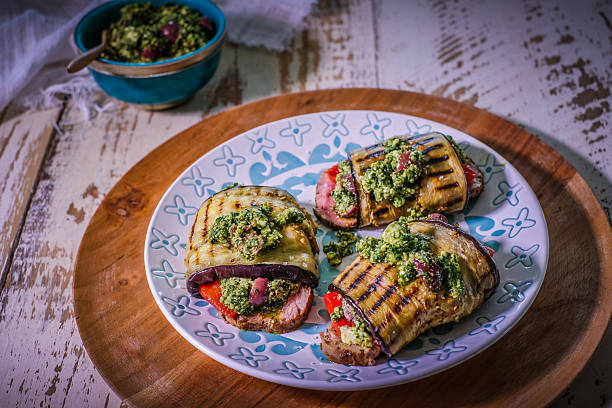 A delightful serving of grilled eggplant slices rolled around a filling of roasted vegetables, garnished with a generous portion of fresh pesto. Served on a patterned plate, this dish presents a visually appealing and delicious vegetarian meal. Perfect for Mediterranean cuisine lovers and healthy eating enthusiasts.