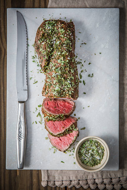 Deliciously prepared beef tenderloin coated with fresh herbs and sliced for serving on a marble board. Accompanied by a small bowl of seasoning, this visually appealing dish combines culinary artistry with mouthwatering presentation. Perfect for gourmet meal inspiration or culinary-related purposes.