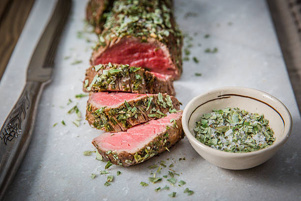 Juicy, herb-crusted roast beef sliced and presented on a marble surface. The vibrant red of the medium-cooked meat contrasts beautifully with the fresh green herbs and the accompanying coarse salt bowl. Ideal for culinary presentations, this image captures the elegance of fine dining and gourmet preparation in a rustic, yet refined, tabletop setting.