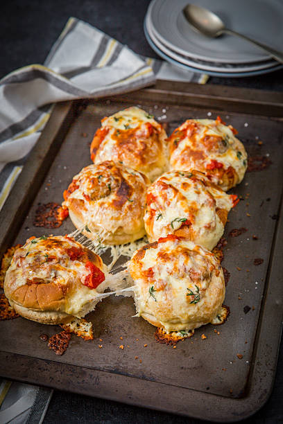 Golden baked rolls topped with melted cheese, garlic, and herbs are showcased on a dark baking tray. The image emphasizes the warm and appetizing appeal of freshly baked bread, perfect for food presentations or recipe illustrations. A contemporary kitchen setup enhances the scene, highlighting homemade delicacies.