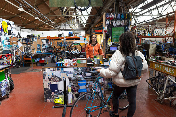 A woman stands at the counter of a bicycle repair and sales shop, speaking with a friendly staff member behind the desk. The shop is filled with bikes, tools, and cycling accessories, creating a busy and welcoming atmosphere. Perfect image to represent local businesses, customer service, sustainable transport, or community-focused repair shops.Videos are available similar to this scenario.