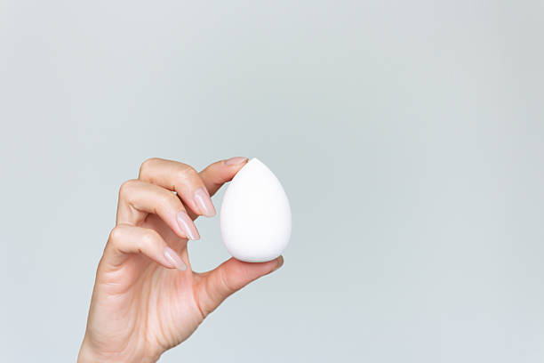 istock A woman's hand holding a makeup sponge for applying foundation on her face, against a neutral gray background. 2234683279