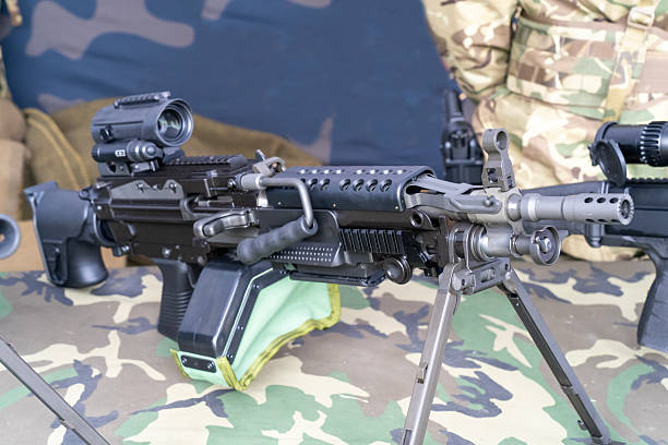 Close-up view of a modern light machine gun with mounted optics, bipod, and ammunition box. The firearm is placed on a camouflage-patterned surface, with soldiers in military uniform visible in the background. The detailed focus highlights the weapon’s mechanical design and tactical use.