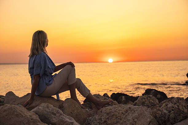 istock Middle-aged woman sitting on coastal rocks at sunset 2234689831