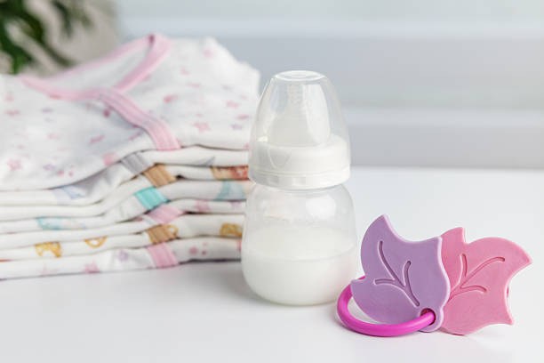 Baby bottle filled with milk beside a pink leaf-shaped pacifier and a stack of folded clothes on a white table.