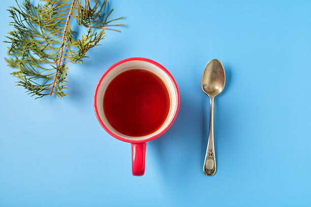 A red cup filled with red tea lies next to a silver spoon on a blue surface. Fresh green foliage enhances the vibrant color scheme of the arrangement.