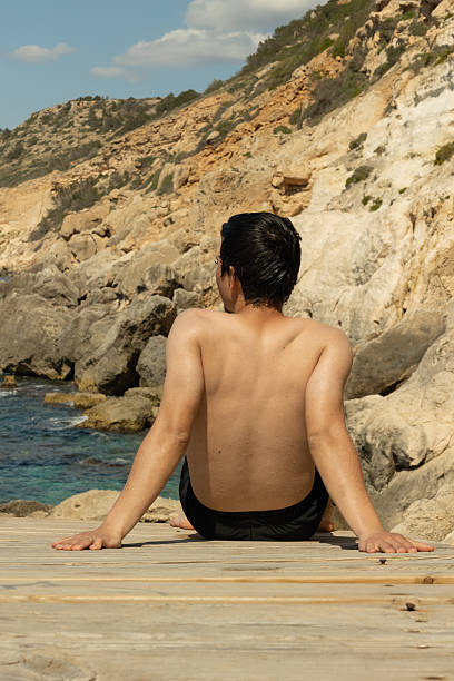 Man sitting on a coastal path after swimming, sunbathing and observing the natural landscape, concept of relaxation, vacation, tourism, and connection with nature