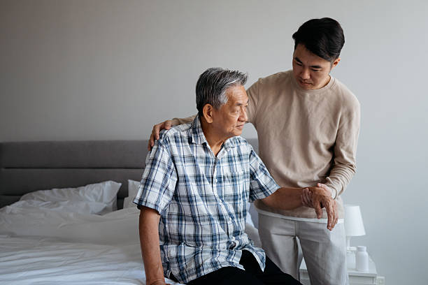 A middle-aged man provides gentle support to his senior father in a bedroom setting, symbolizing compassion, family connection, and caregiving.