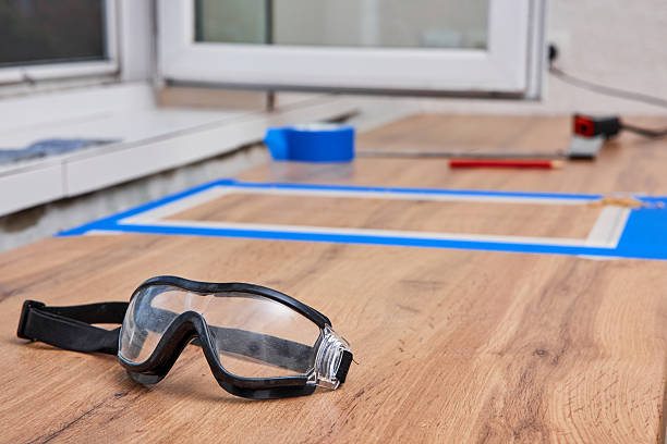 Protective safety glasses rest on an outdoor table surface, illuminated by a warm sunlit background with soft-focus natural scenery. Represents safety, outdoor work, and the sunny environment.