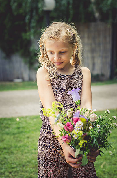 A young girl with curly blonde hair kneels in a meadow, picking wildflowers. She wears a blue blouse and holds a bouquet of colorful flowers.