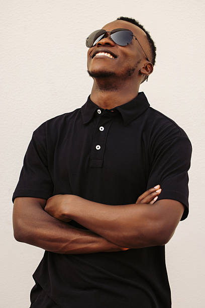 A cheerful individual dressed in a black shirt and sunglasses smiles confidently, standing against a simplistic cream background. The image portrays a mood of positivity, modern style, and confidence. Perfect for illustrating happiness, fashion, or a casual theme in projects and advertisements.