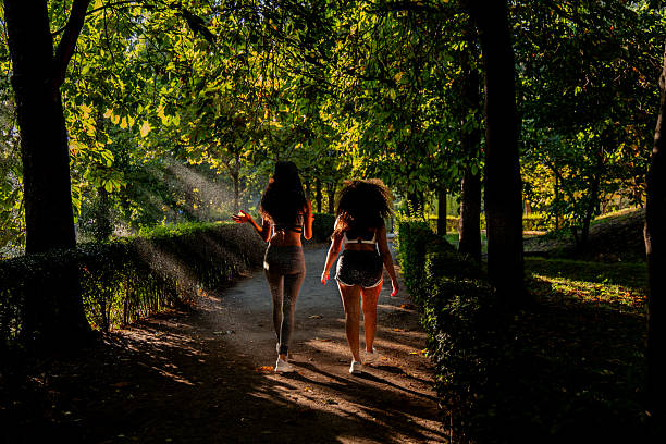Two young latin women, dressed in sportswear, are walking in a park during a sunny morning, enjoying the fresh air and the light filtering through the trees