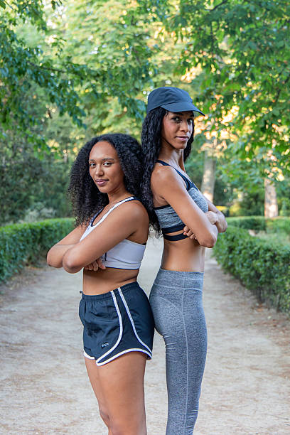 Two smiling Latin sportswomen standing back to back in a park, crossing their arms confidently while wearing stylish sportswear. Enjoying the summer day, they embody determination and friendship