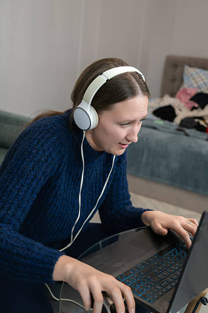 istock Young woman wearing headphones is working or studying from home, using laptop and mouse, concentrating on the screen while sitting in her messy bedroom. Vertical photo 2234729697