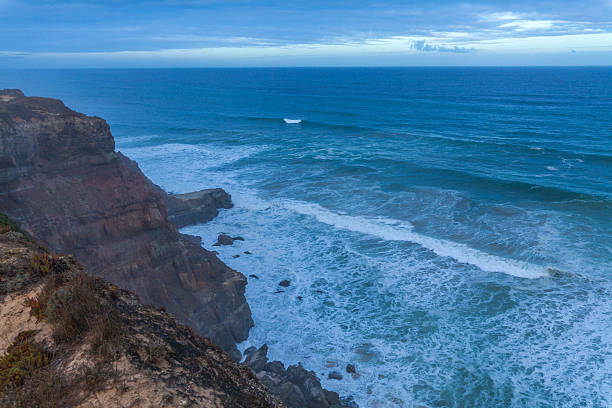 A dramatic coastal cliff overlooks powerful ocean waves crashing against rocky shores under a moody blue sky with scattered clouds