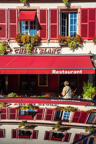 Ribeauville, France - August 24, 2025: A charming restaurant facade with red shutters and flowers, reflecting on the roof of a car, featuring a woman walking by
