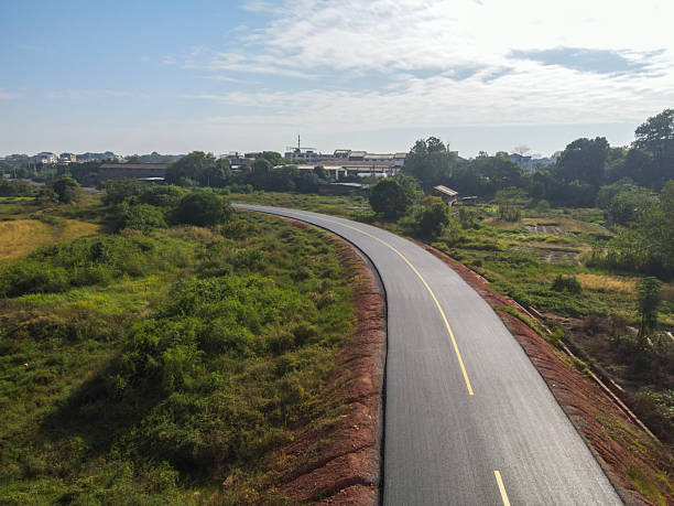 A breathtaking aerial view captures a smooth asphalt road winding through a dense, sun-drenched forest. Golden sunrise light filters through the trees, highlighting the road's sleek texture and creating long, dramatic shadows. The scene feels both serene and powerful, offering a high-quality, minimalist backdrop perfect for an automotive advertisement—where nature meets precision engineering.