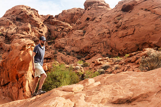This is a photograph of a man in his 40s wearing a baseball hat while hiking on red rocks in the Nevada desert on a summer day.