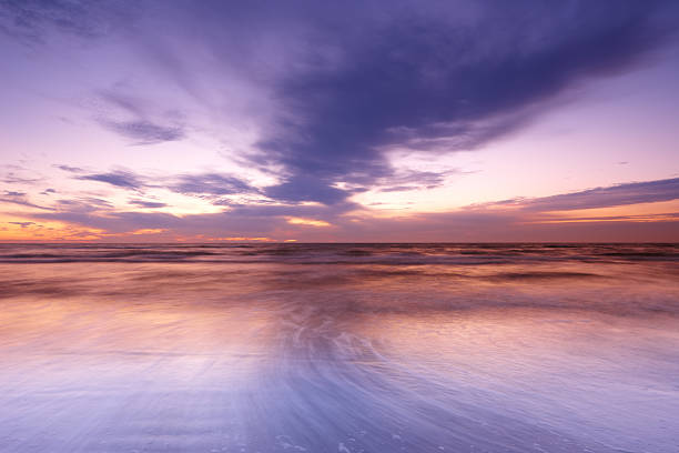 A captivating scene of a beach during twilight, where vibrant purple and orange colors fill the sky, reflecting elegantly on the water. The gentle waves create a serene ambiance, ideal for relaxation and inspiration.