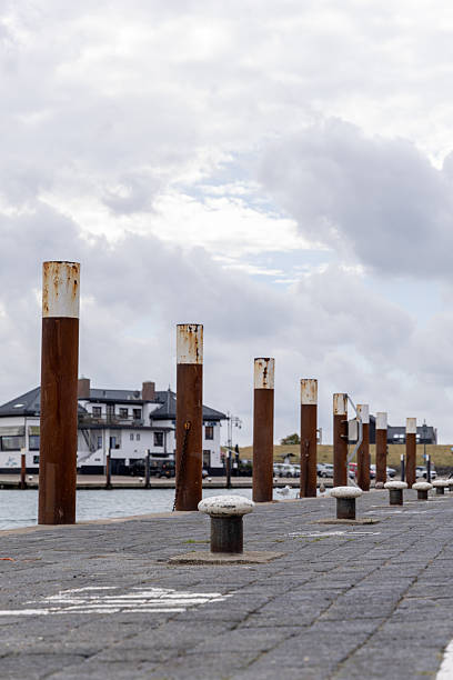 Weathered wooden posts line a tranquil waterfront, with a distant building and cloudy sky creating a serene atmosphere, perfect for coastal lifestyle themes and nautical imagery