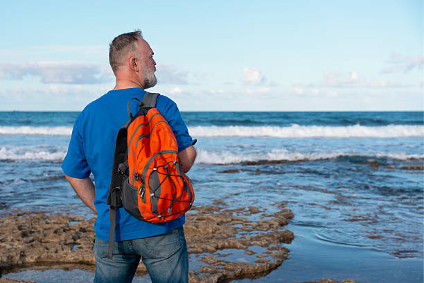 A man stands on a rocky shoreline, looking out at the ocean waves. He wears a blue shirt and an orange backpack, with a bright sky overhead.