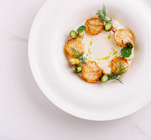 A fine dining dish features beautifully seared scallops arranged in a circular pattern on a white plate. Fresh herbs garnish the dish, complementing the aromatic sauce.