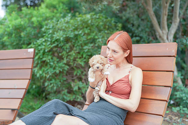 A woman is sitting on a bench with a small dog in her lap. The dog is brown and white. The woman is wearing a red top and a black skirt. The scene is peaceful and relaxing, with the woman