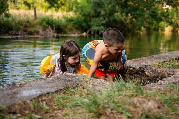 Vertical photo of two children sitting on a small wooden bridge near a river, surrounded by tall grass and rocks. Peaceful summer outdoor scene showing childhood and nature connection.
