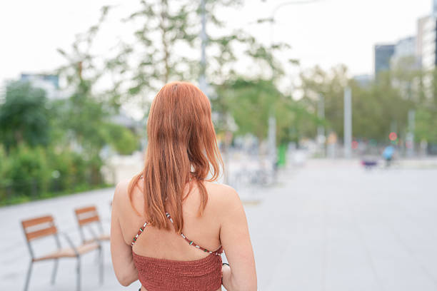 A woman with red hair is walking down a sidewalk. She is wearing a brown dress and a brown shirt. There are several chairs and benches along the sidewalk, and a few people are walking by