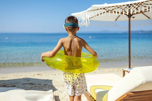 A young child stands on a sunny beach holding an inflatable yellow float, with a serene sea view. The atmosphere evokes the feeling of relaxation and the joy of summer vacation.