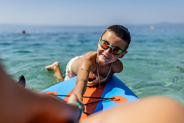 A cheerful boy wearing goggles having fun while lying on a paddleboard in pristine, crystal-clear water under a sunny sky, capturing the essence of summer joy and outdoor adventure.