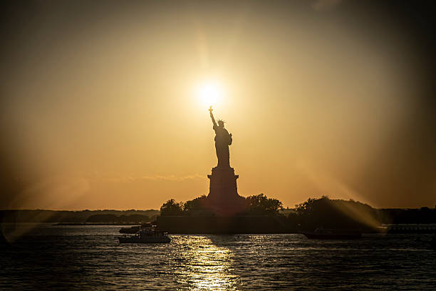The Statue of Liberty in New York silhouetted against a golden sunset sky over the Hudson River.