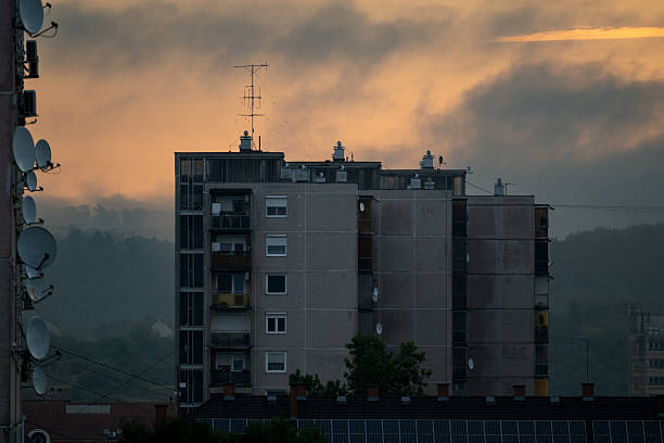 A high-rise apartment building is seen in silhouette against a dramatic sunset sky with orange and gray clouds. Satellite dishes line the side of a neighboring structure, while misty hills fade into the background, creating a moody and atmospheric urban scene.