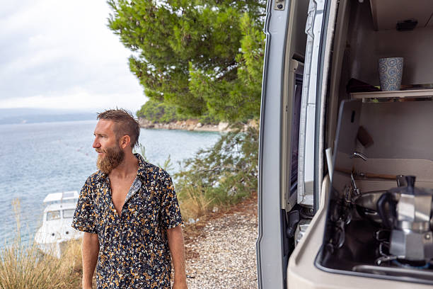 Bearded man in floral shirt standing by camper van on rocky coast. Relaxed adult male appreciating ocean scenery and nature. Outdoor lifestyle and travel concept.