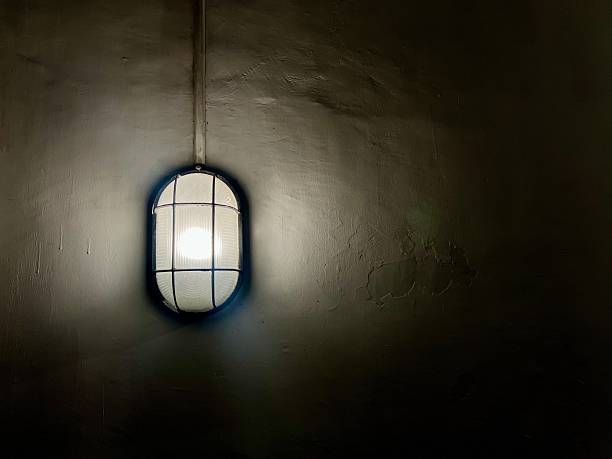 A wall-mounted industrial light fixture with a protective metal cage and frosted glass cover, emitting a soft glow against a textured wall.