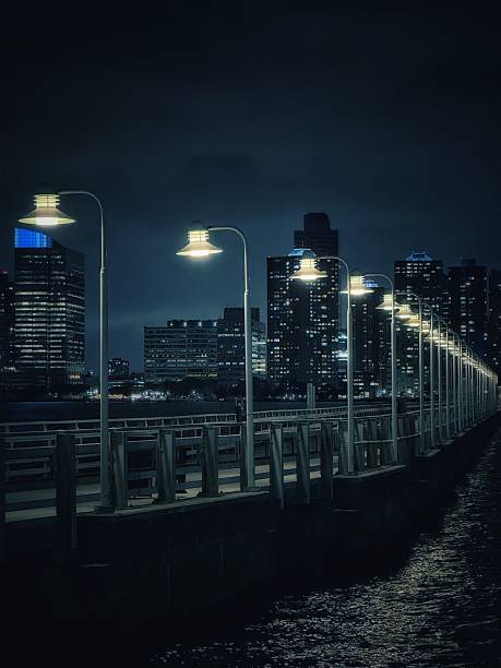 New York City, USA – A row of illuminated street lamps on a pier along the waterfront at night, with the skyline of New Jersey visible in the background.