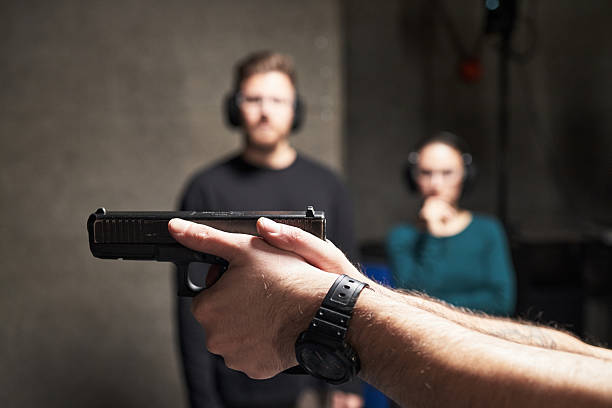 Caucasian young adult man holding handgun at shooting range with blurred Caucasian young adult man and woman standing in background wearing protective earmuffs observing