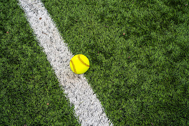 A vibrant neon yellow softball rests directly on a thick, white-painted boundary line, creating a strong visual contrast against the lush green artificial turf. The detailed texture of both the synthetic grass and the ball's red stitching is clearly visible, grounding the image in the tangible world of sport.