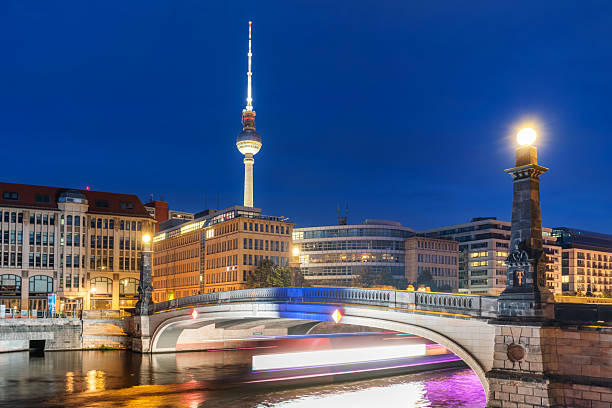 Nighttime cityscape of Berlin showcasing a brightly illuminated bridge, water reflections, and the iconic television tower with surrounding modern architecture under a deep blue evening sky.
