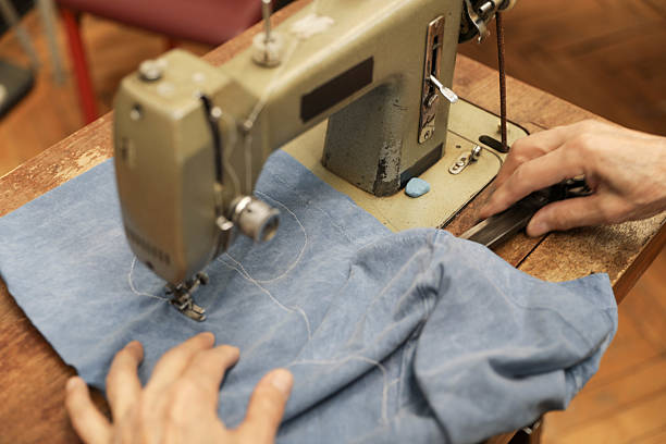 Back View Of A Senior Woman (80+) In Glasses Operating A Sewing Machine, Hands Guiding Blue Fabric On A Wooden Worktable Inside A Home Sewing Workshop, Illustrating Craftsmanship, Needlework Skills, DIY Creativity, And Active Aging.