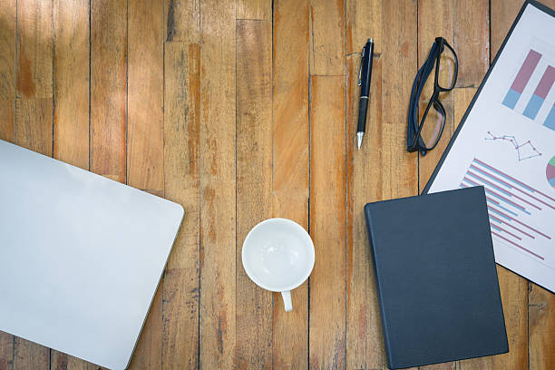 Empty cup of coffee, laptop, notebook, pen, glasses and graph on wooden table. Top view with copy space.