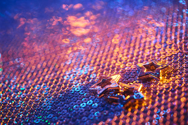 A festive still life showcasing metallic star-shaped Christmas decorations arranged on a glowing sequin surface. Blue and orange neon lighting creates a dreamy, celebratory mood, perfect for holiday and New Year themed visuals.