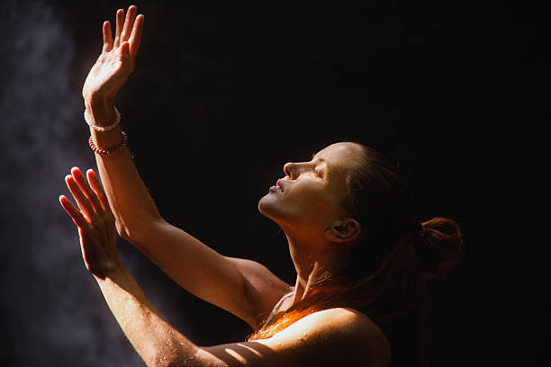 A woman with her face illuminated by sunlight, raising her arms gracefully against a darkened backdrop. The composition emphasizes calmness, spirituality, and connection with light and nature.